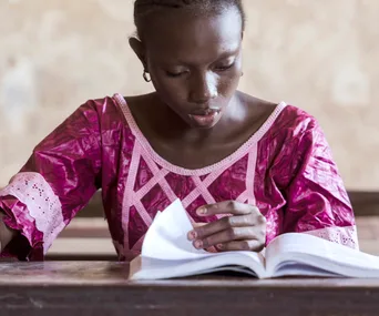 Young girl in a pink dress reading a book attentively at a desk.