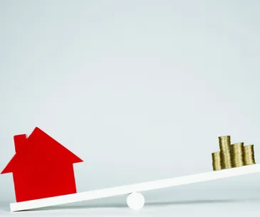 Seesaw with red house model on one end and stack of coins on the other, balancing concepts of property and investment.