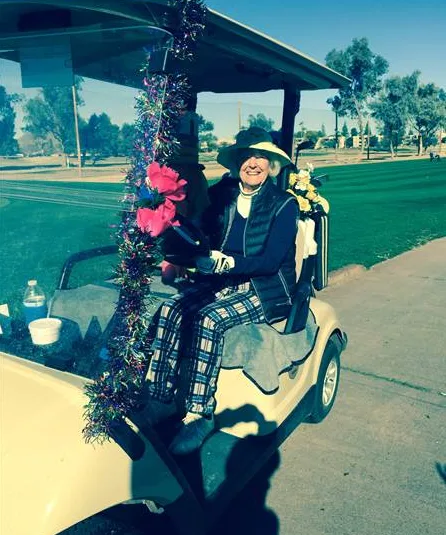Golfer in a decorated golf cart on a sunny course, wearing a wide-brimmed hat and plaid pants, smiling warmly.