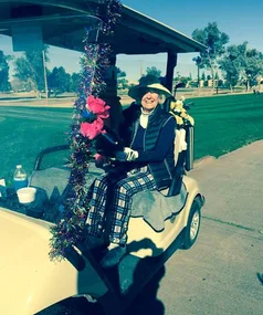 Golfer in a decorated golf cart on a sunny course, wearing a wide-brimmed hat and plaid pants, smiling warmly.