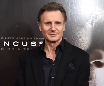 A man in a black suit stands in front of a film poster backdrop.