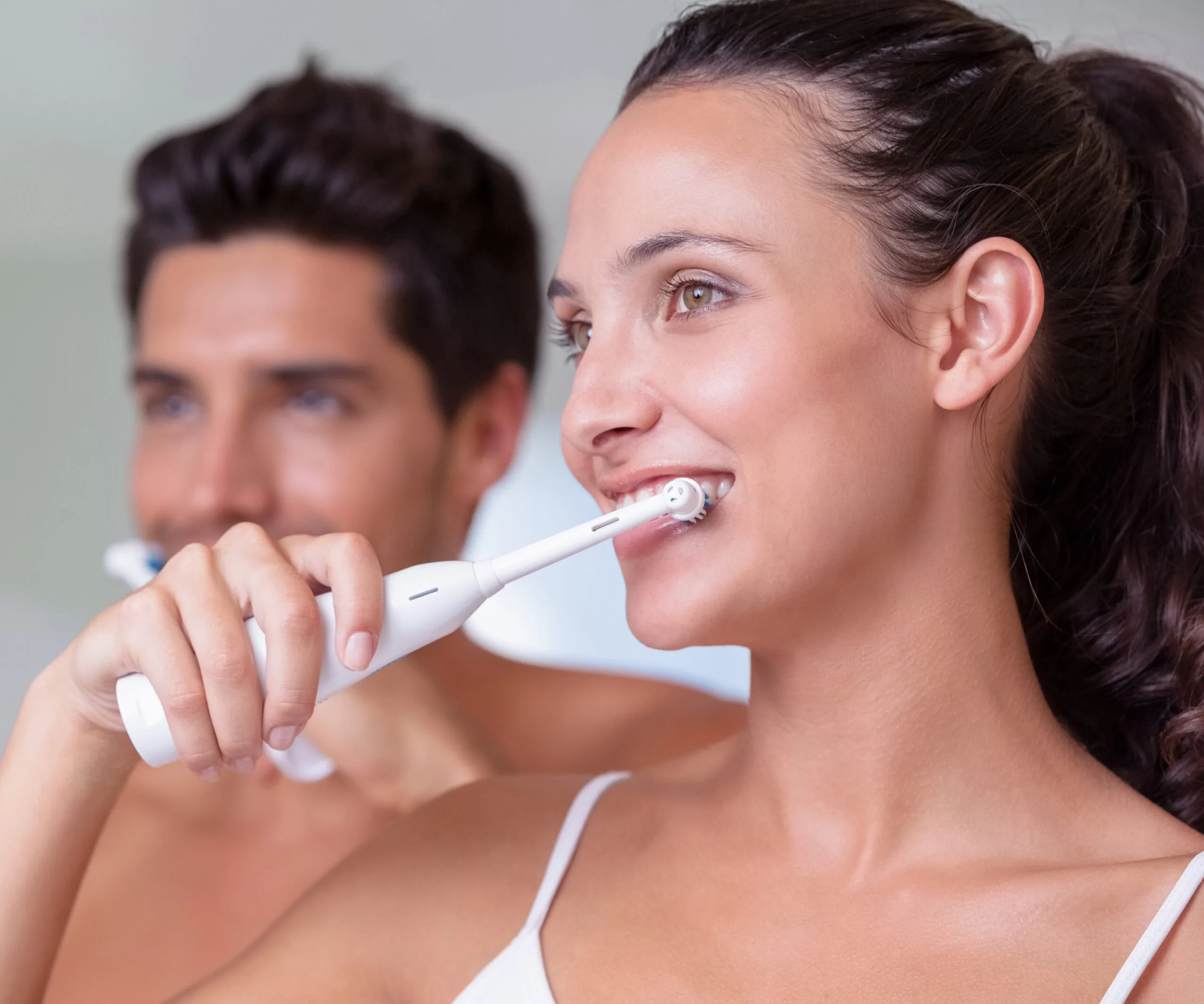 Two people brushing their teeth with electric toothbrushes, smiling and looking in a mirror.