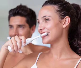 Two people brushing their teeth with electric toothbrushes, smiling and looking in a mirror.