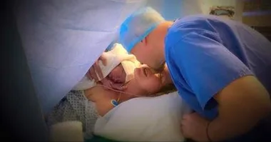 Mother holding newborn in hospital bed, partner leaning in, during caesarean birth.