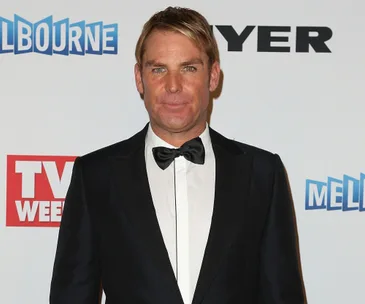 A man in a tuxedo stands in front of a white backdrop with logos, facing the camera.