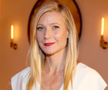 A person with long blonde hair and red lipstick in a white outfit stands indoors, with soft lighting and sconces in the background.