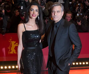 A couple poses on a red carpet; the woman wears a sequined black dress, and the man is in a black suit. Photographers are behind.