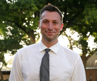 Man in white shirt and gray tie, smiling outdoors with trees blurred in the background.