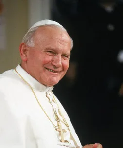 Smiling elderly man in religious attire with a cross necklace and white cap.