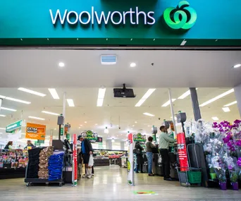 Entrance of a Woolworths store with customers entering, visible signage, and display of flowers and products inside.