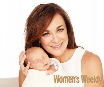 Woman smiling, holding a newborn wrapped in a white blanket, featured in The Australian Women's Weekly.