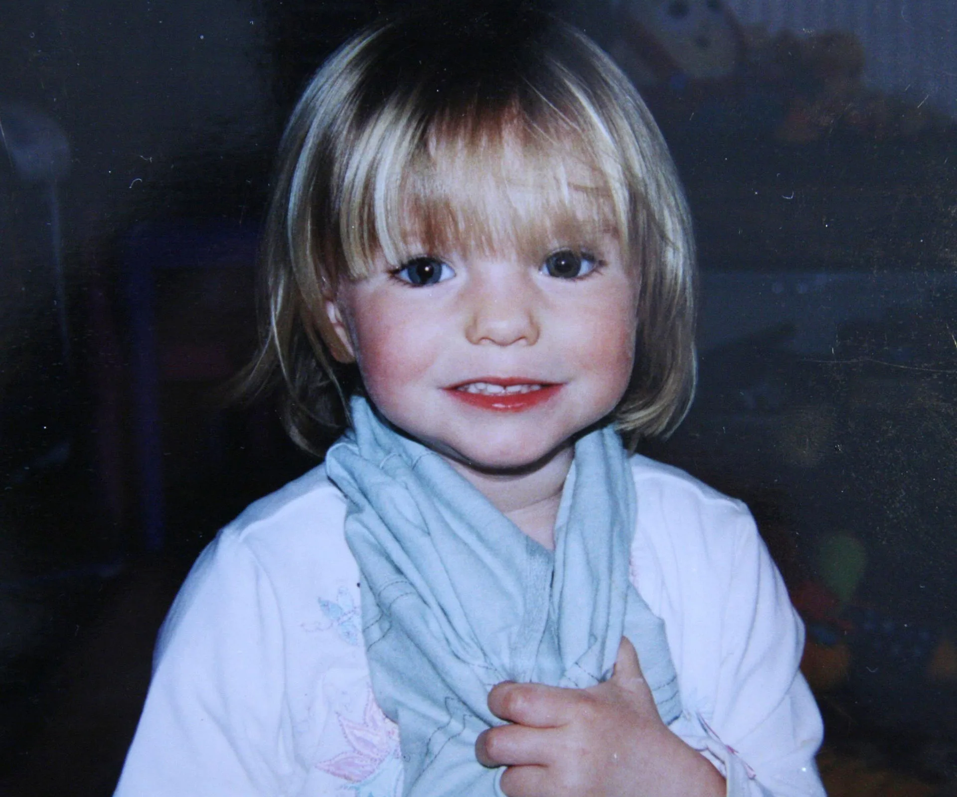 A young child with short blonde hair smiles, holding a light blue scarf against a dark background.