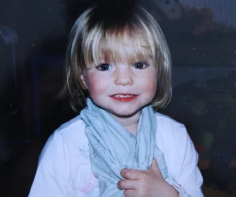 A young child with short blonde hair smiles, holding a light blue scarf against a dark background.