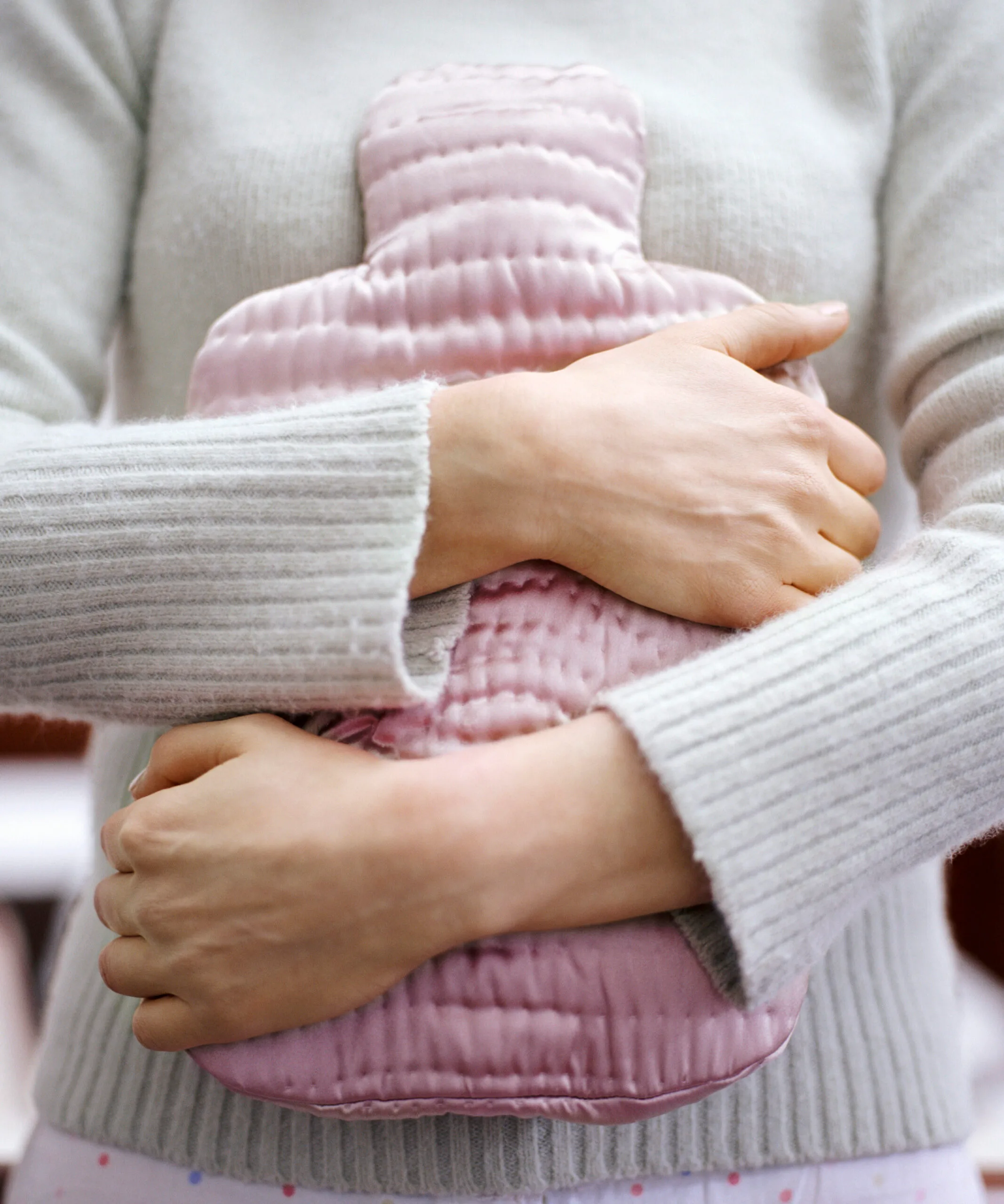 A woman in a grey sweater holds a pink hot water bottle to her abdomen.