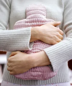 A woman in a grey sweater holds a pink hot water bottle to her abdomen.