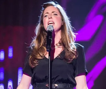 Singer performing passionately on stage with a microphone, wearing a black outfit, colorful lights in the background.