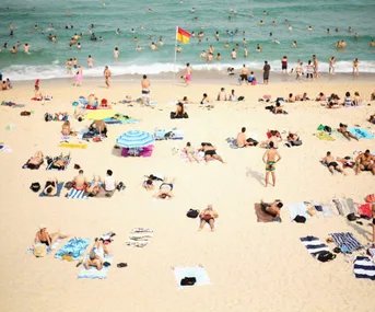 Beach scene with people sunbathing on sand, umbrellas, towels, and swimmers in the ocean, under a sunny sky.