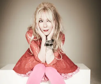 Morwenna Banks in a red dress and pink tights, sitting with hands on cheeks, looking playfully to the side.