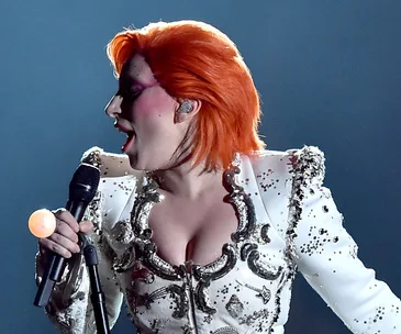 Singer with bright orange hair in a sequined white outfit performing on stage, holding a microphone.
