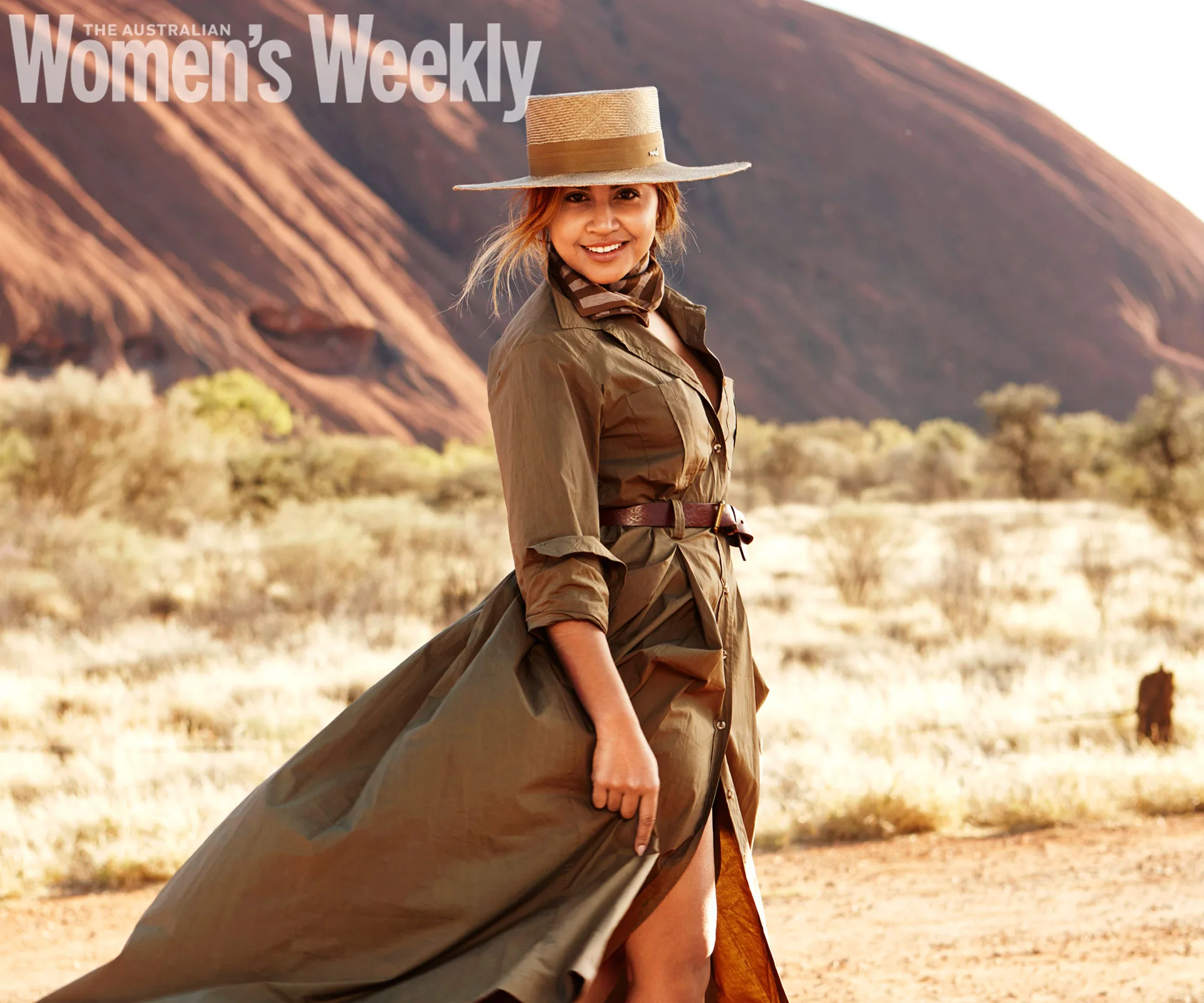 Woman in a flowing dress and hat stands in a desert landscape, smiling, with a rocky backdrop.