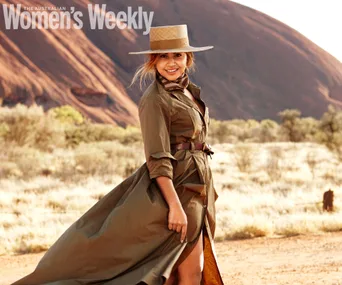 Woman in a flowing dress and hat stands in a desert landscape, smiling, with a rocky backdrop.