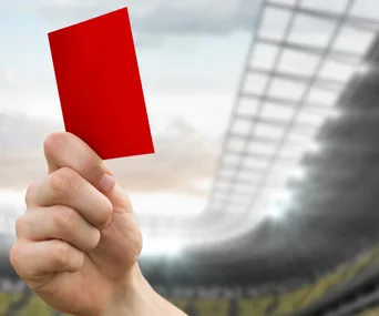 A referee's hand holding a red card on a football field background.