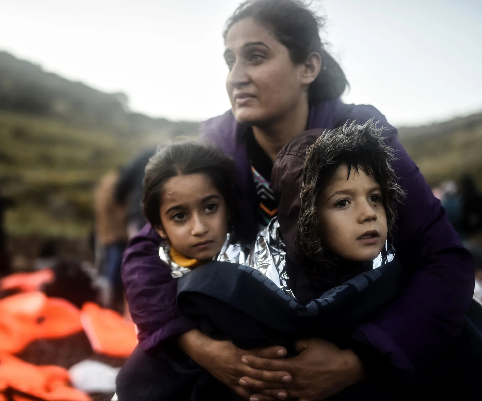 A woman holding two children wrapped in blankets, sitting outdoors with a blurred background.