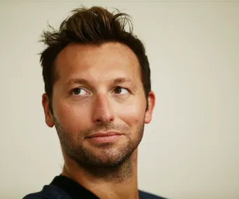 A person with short hair and stubble looks slightly up and to the side, against a neutral background.
