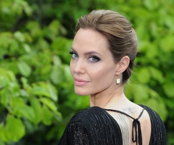 I don’t know who this person is, but they are posing outdoors in elegant attire, with greenery in the background.