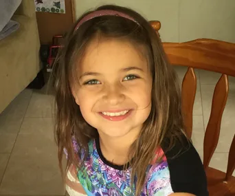 A young girl with long hair smiles brightly, wearing a colorful shirt and headband, sitting on a wooden chair indoors.