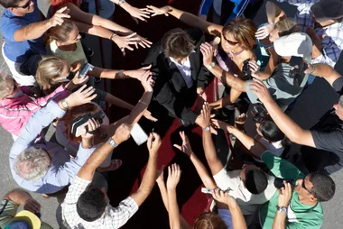 Crowd surrounds a person in a suit on a red carpet, reaching with cameras and pens for autographs.