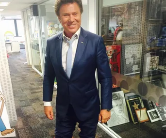 Man in a blue suit, smiling, standing in an office corridor with framed photos and memorabilia on the walls.