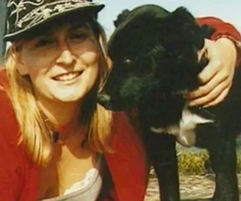 A person in a red sweater and cap smiling next to a black dog outdoors.