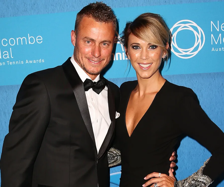 A couple in formal attire smiles at a tennis awards event with a blue background.