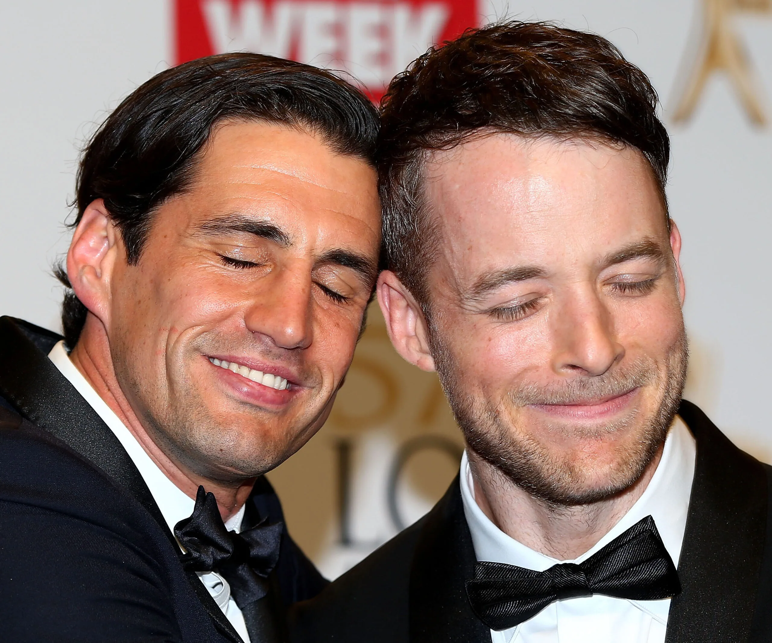 Two men in black tuxedos with eyes closed, smiling closely together at a formal event.