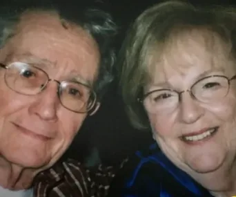 Elderly couple smiling for a photo, both wearing glasses, close together in a warm moment.