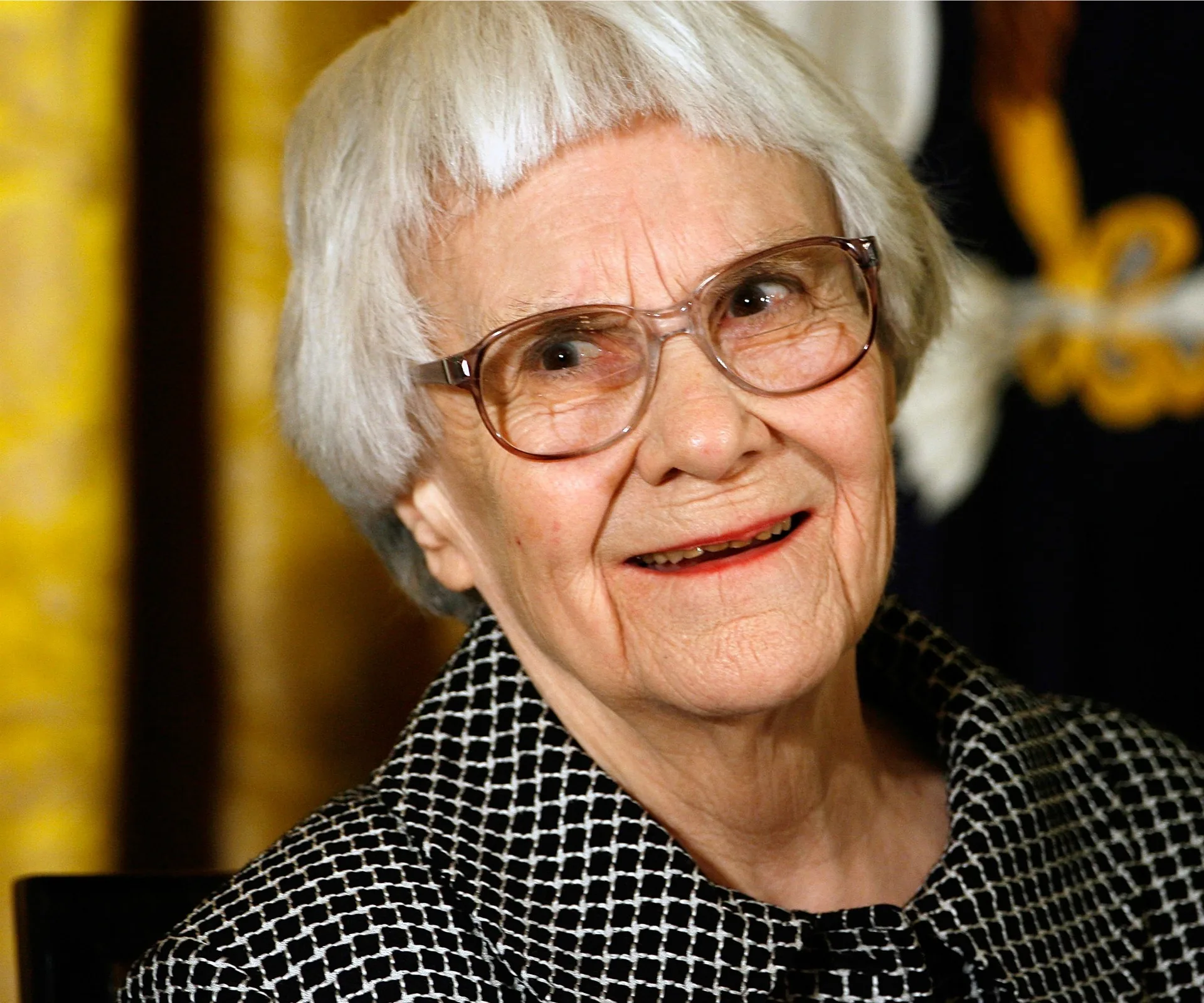Elderly woman with short white hair and glasses, smiling, in a checkered outfit.