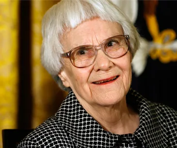 Elderly woman with short white hair and glasses, smiling, in a checkered outfit.