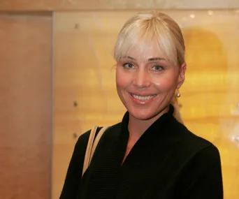 A person with blonde hair smiling, wearing a black outfit, and standing against a wooden background.