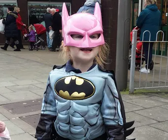 Young girl in a Batman costume with a pink mask, smiling proudly on a busy sidewalk.