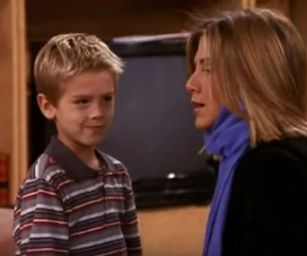 Young boy in a striped shirt stands next to a woman with blonde hair wearing a blue scarf, inside a room.