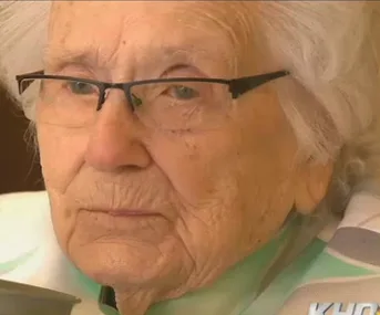 Elderly woman with glasses sitting, looking forward with a neutral expression.