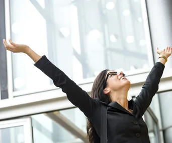 Businesswoman in black suit celebrating with arms raised in front of a modern glass building.