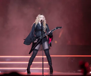 Madonna performing on stage, wearing a black outfit and playing a black guitar.