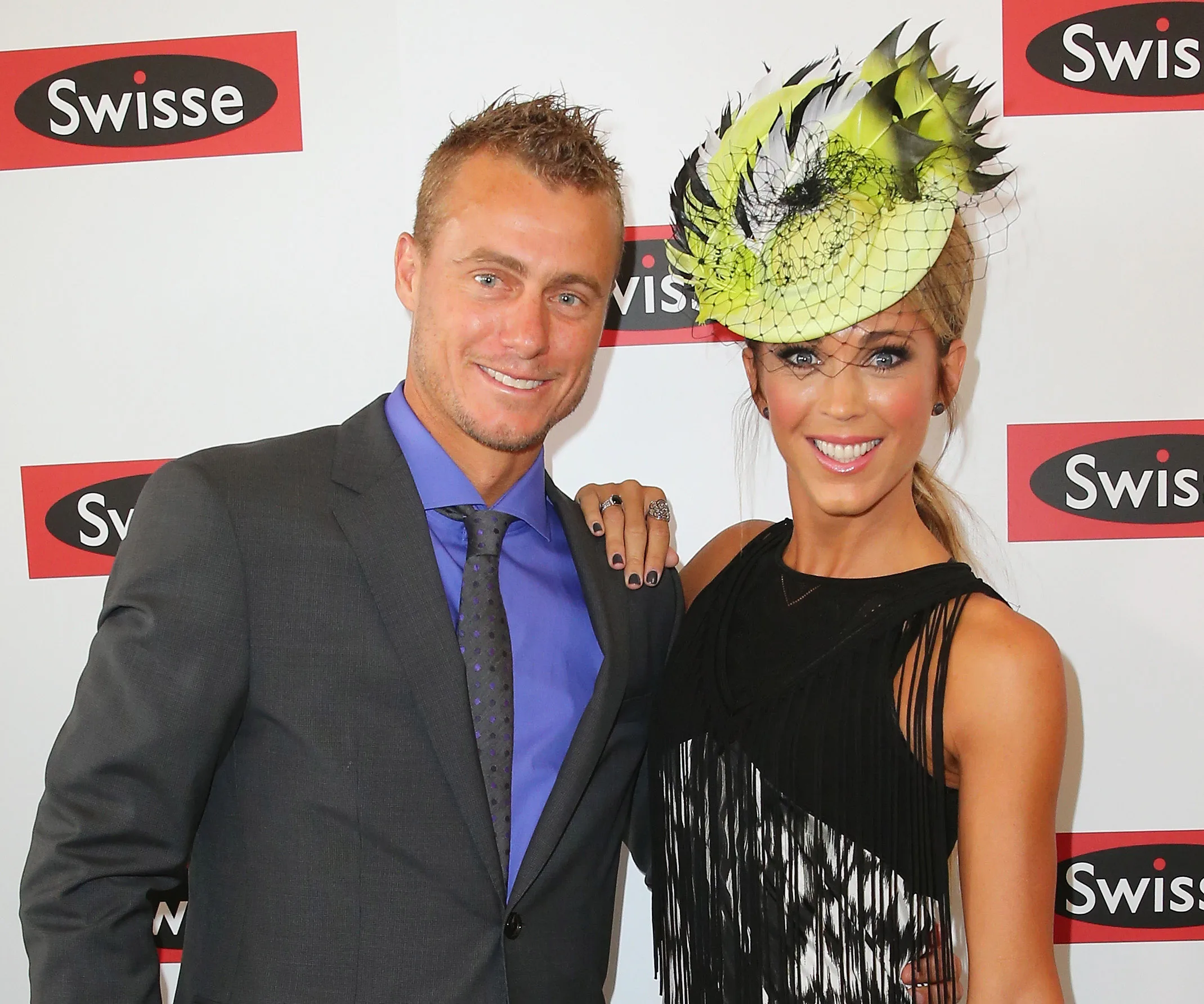 A smiling couple posing on a red carpet, woman wearing a decorative hat.