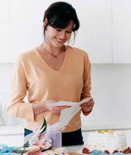Woman in a peach sweater smiles while reading a card, with a cake and flowers on a white kitchen counter.