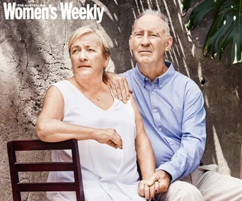 Elderly couple seated, woman in white and man in blue shirt, holding hands and looking pensive; outdoors with a cracked wall.