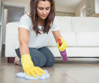 Woman cleaning tile floor with spray and cloth, wearing yellow gloves in a living room near a white sofa.