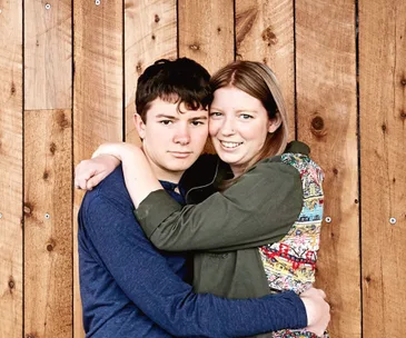 A young woman and teenager hugging in front of a wooden wall, both smiling warmly at the camera.
