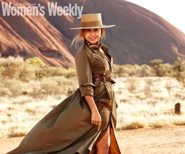Woman in a long coat and hat standing in front of Uluru, smiling with the desert landscape in the background.